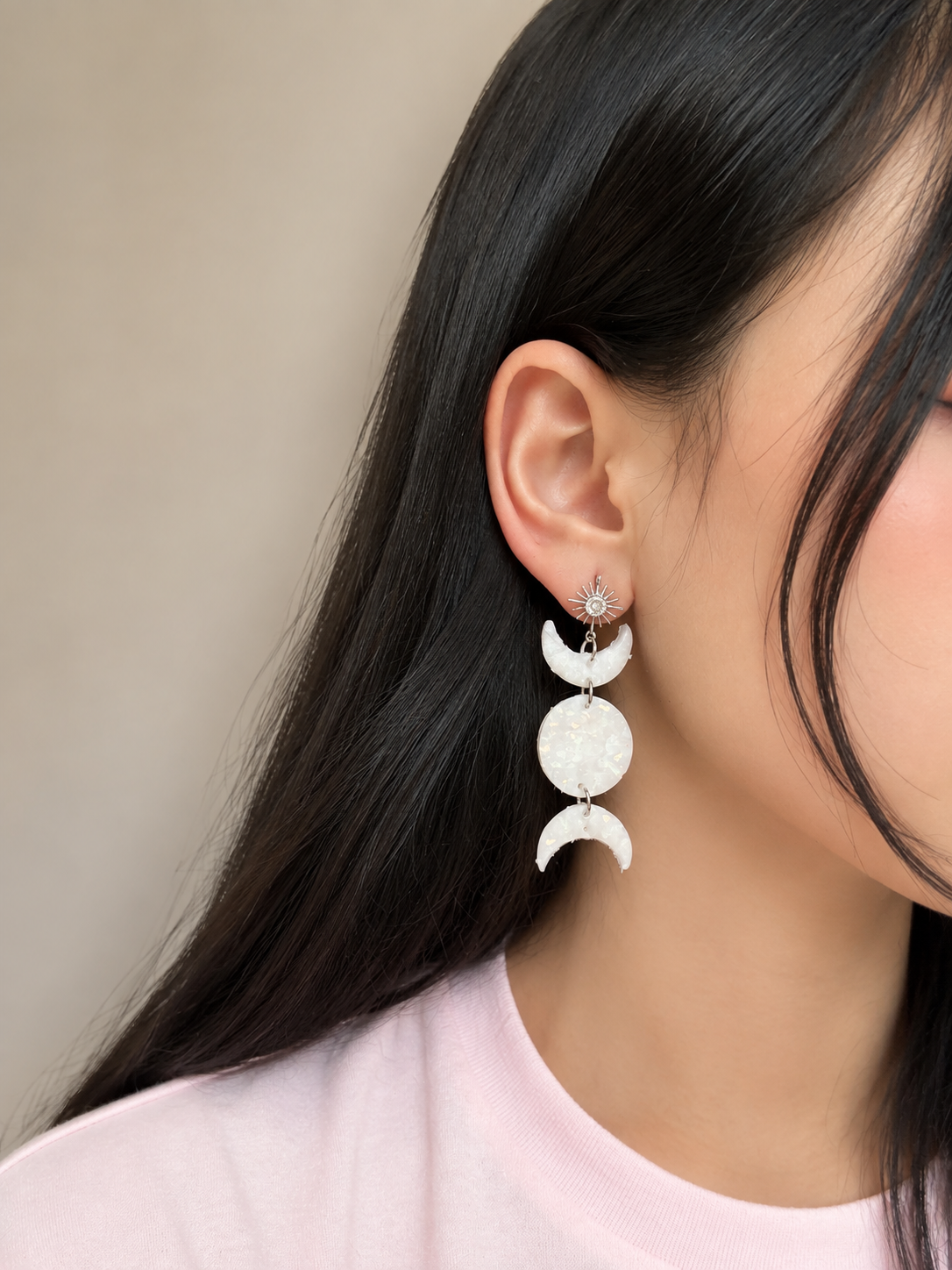 A close-up of a model's ear wearing a dangling triple moon phase earring. The earring features three textured white crescent moon and full moon charms hanging from a silver-toned star stud.