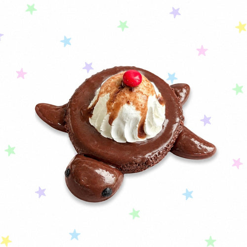 A novelty chocolate turtle-shaped dessert topped with whipped cream, chocolate drizzle, and a red cherry, set against a white background with scattered pastel star graphics.